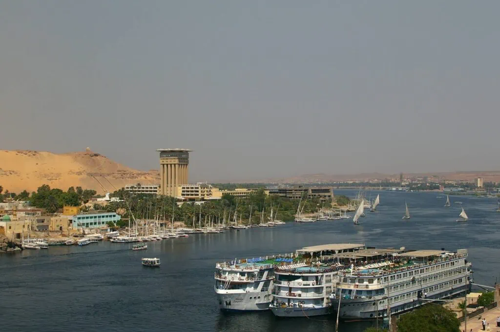Aswan corniche on the east bank of the Nile, with three 5-star Luxury Nile Cruise vessels moored side-by-side at the southern terminus of the standard Luxor–Aswan cruise route, traditional feluccas anchored along the riverbank, the Mövenpick Resort Aswan on Elephantine Island in the distance, and the west bank desert escarpment beyond.