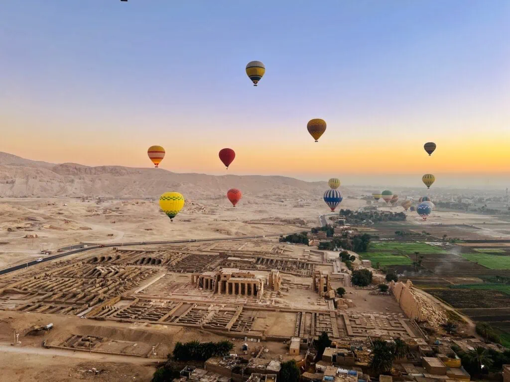 Hot-air balloons at sunrise over the Ramesseum, the mortuary temple of Ramses II on Luxor's West Bank, with the temple's preserved hypostyle hall and mudbrick storage magazines visible from the air.