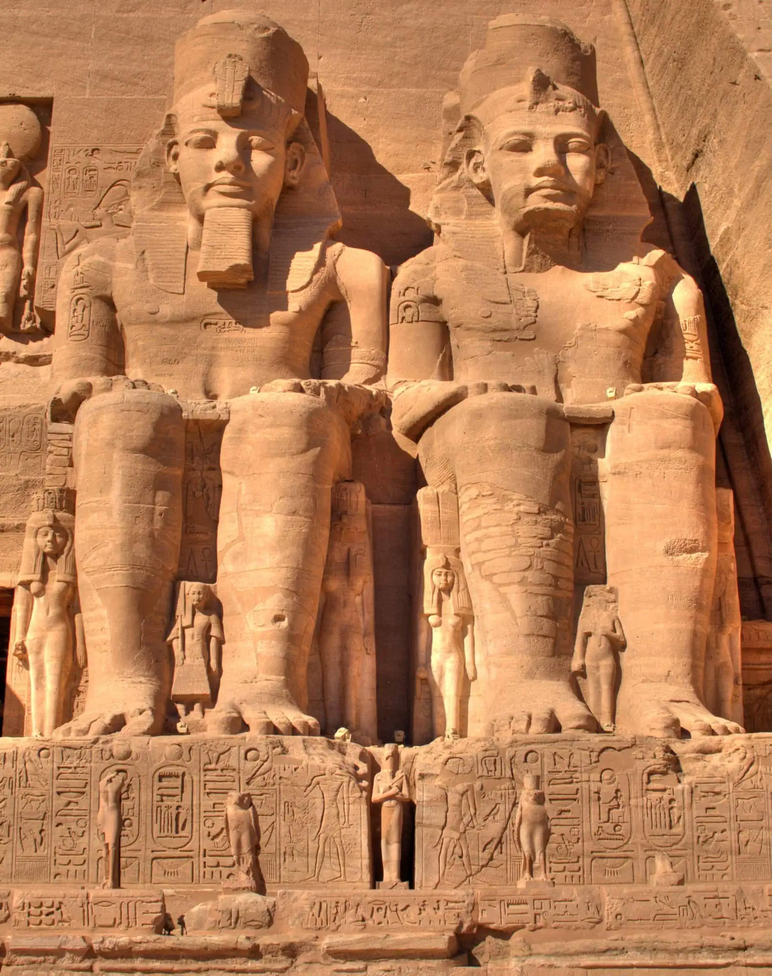 Close view of two of the four giant seated statues of Ramesses II at the facade of the Great Temple of Abu Simbel, with smaller royal family figures at their legs and carved hieroglyphs below, Egypt.