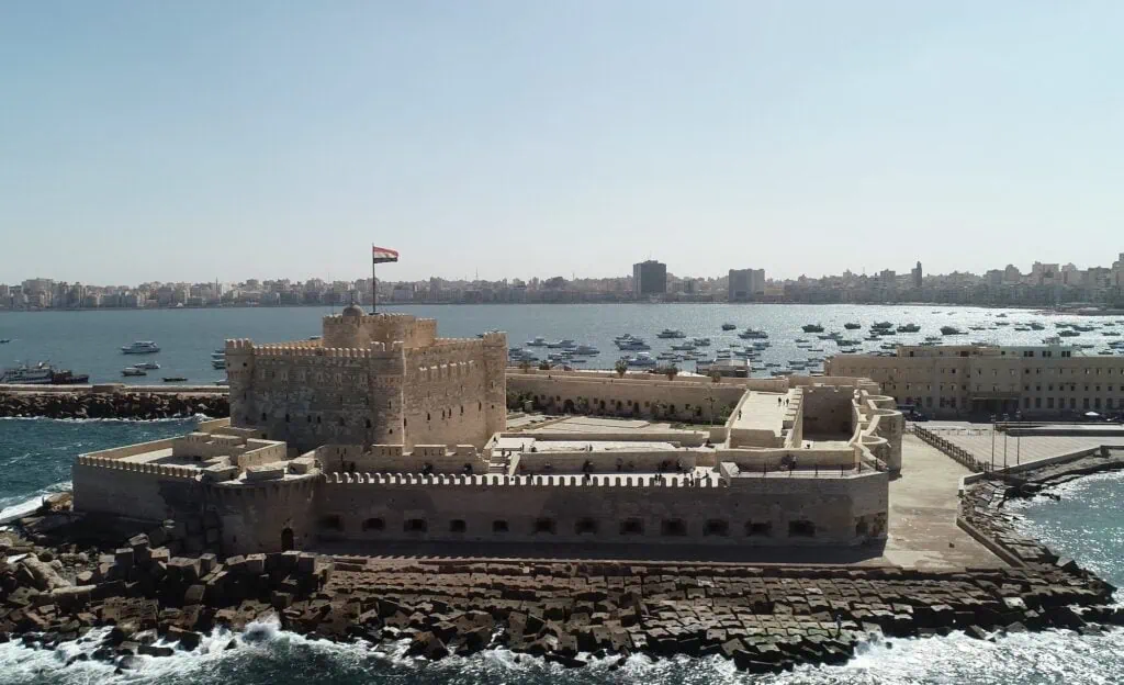 An aerial drone view of the Qaitbay Citadel in Alexandria, Egypt, showing the fortified stone structure standing on a peninsula extending into the Mediterranean Sea, with the modern city of Alexandria visible in the background.