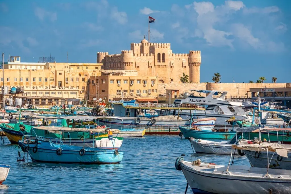 The medieval Qaitbay Citadel rising from a stone breakwater at the entrance to Alexandria's harbor, with the calm Mediterranean Sea in the foreground under a clear blue sky.