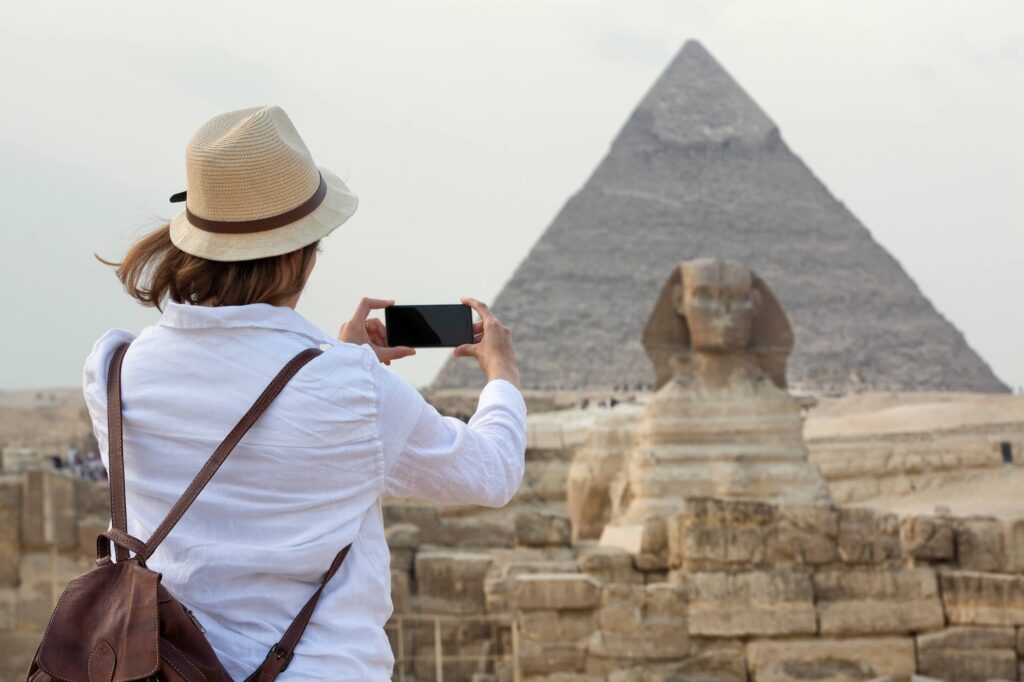 A young woman taking a photograph in front of the Great Sphinx of Giza with the pyramids in the background, Cairo, Egypt.