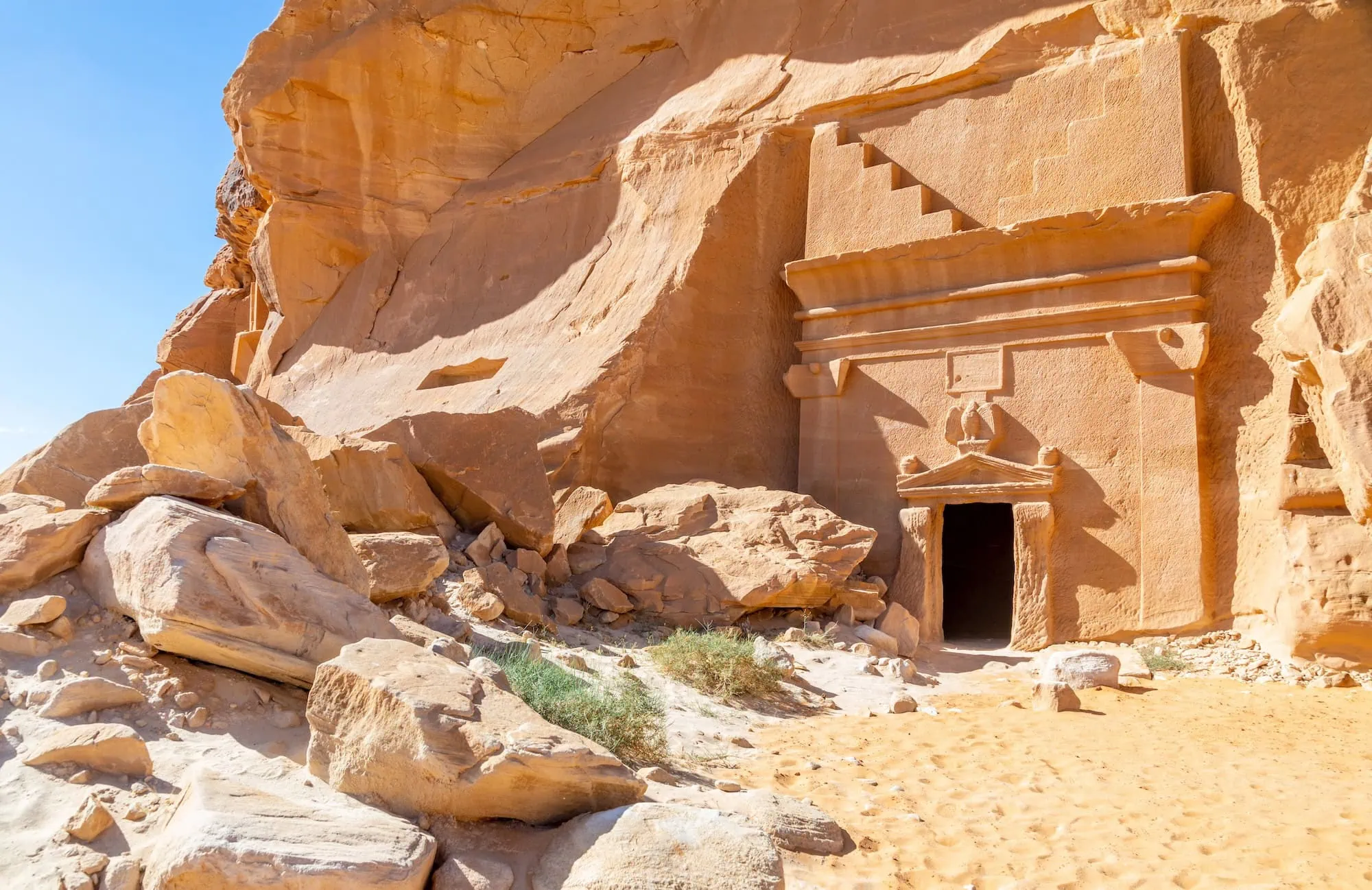 Carved Nabataean rock-cut tomb facade at Jabal al-Banat in Hegra archaeological site, AlUla, Saudi Arabia — single-chamber tomb with classical pediment, urn finials, and stepped crown