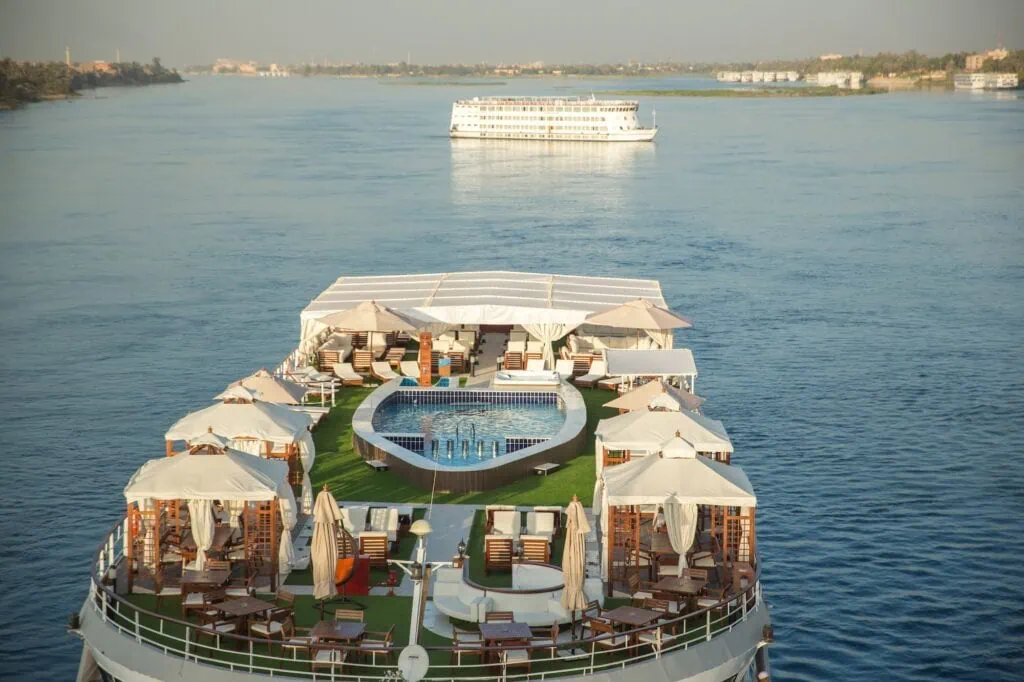 The aft sun deck of a 5-star Luxury Nile Cruise vessel with central pool, white-draped cabañas, sun loungers on artificial grass, and another cruise vessel visible on the Nile behind. 5-Day Nile Cruises