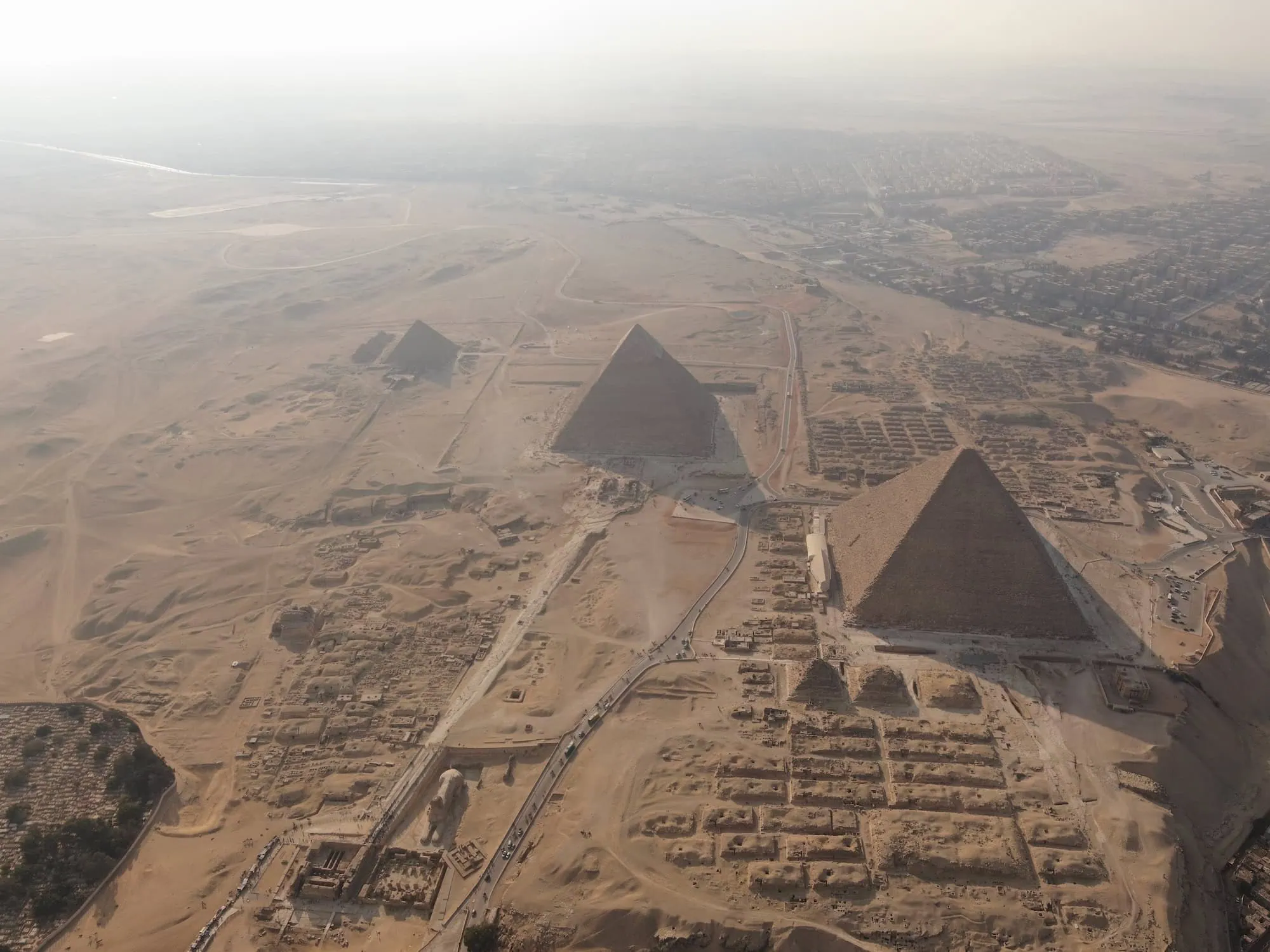 An aerial view of the Giza pyramid complex showing the three main pyramids of Khufu, Khafre, and Menkaure rising from the desert plateau, with surrounding mortuary structures and the modern Cairo suburbs visible at the eastern edge.