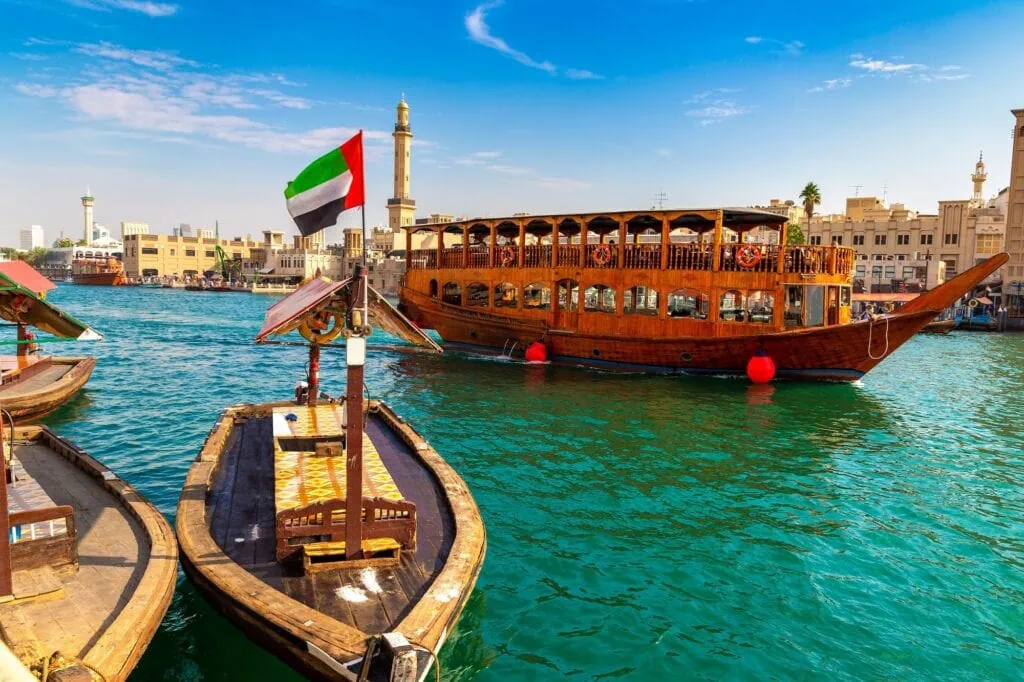 Dubai Creek Cruise – traditional wooden dhow sailing past the Grand Bur Dubai Masjid Mosque along the bay in Dubai, UAE