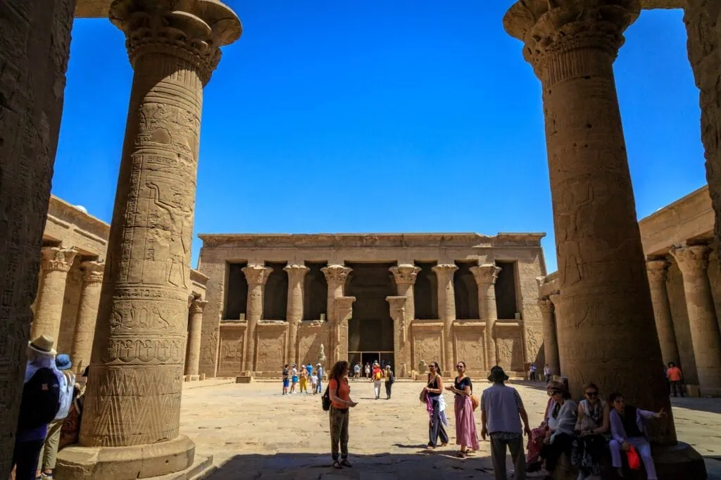 Cruise passengers on a small-group shore excursion exploring the peristyle courtyard of the Temple of Horus at Edfu, with massive Ptolemaic columns covered in hieroglyphic reliefs in the foreground.