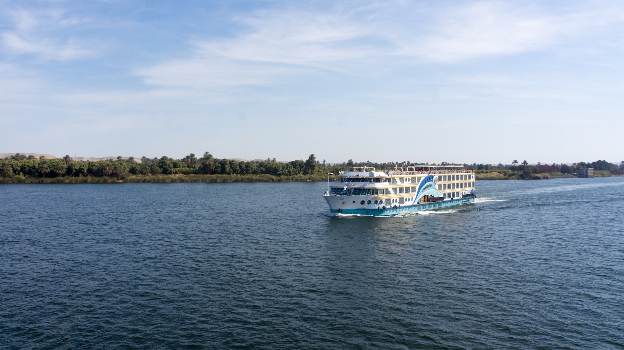 A white and blue Nile cruise vessel sailing on calm water between Luxor and Aswan, with lush green palm groves and cultivated riverbanks visible in the foreground.