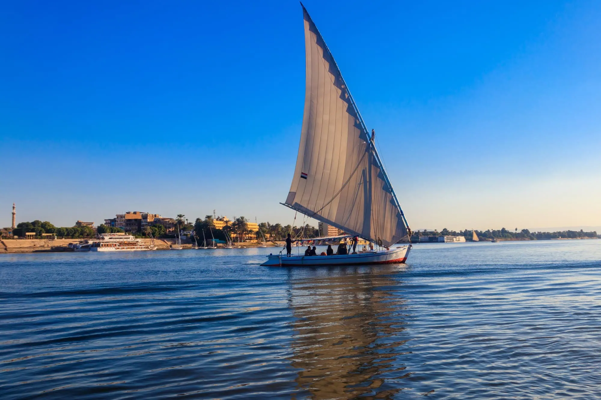 A traditional felucca sailboat with a triangular lateen sail gliding on the calm waters of the Nile River at sunset, with the sky glowing in warm tones, Egypt.