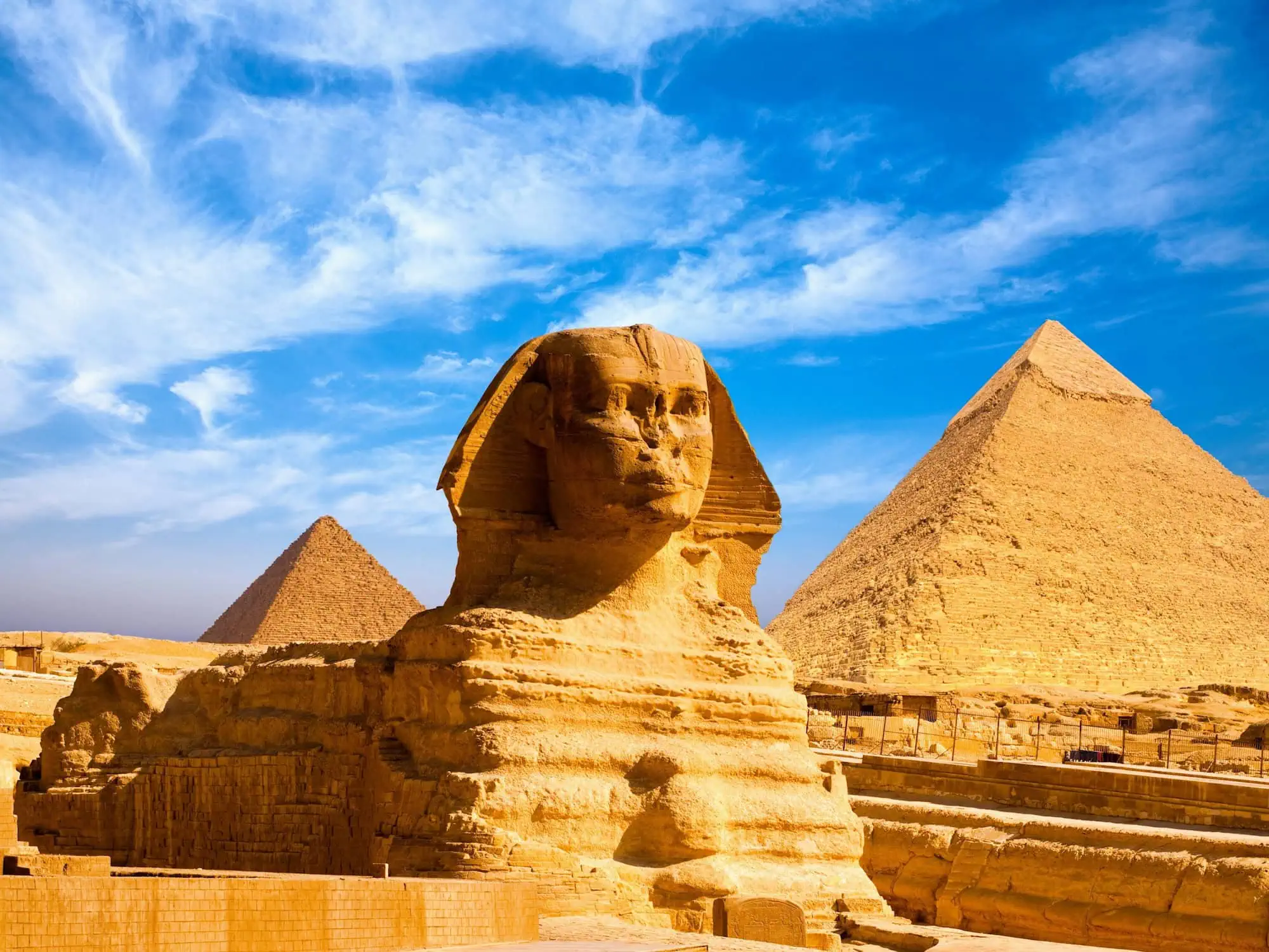 The Great Sphinx in profile at the foot of the Giza plateau, with the pyramids of Khafre and Menkaure rising behind on the western desert outside Cairo - Classic Egypt Tours