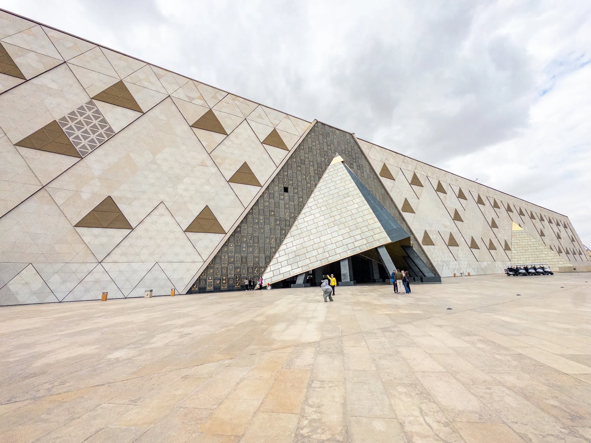 Modern Grand Egyptian Museum building with geometric triangular facade and visitors at entrance plaza