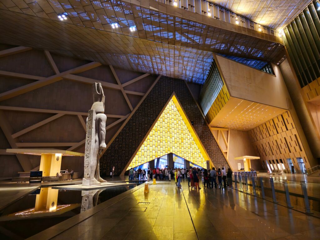 Grand atrium of the Grand Egyptian Museum at Giza with the colossal Ramses II statue rising at the foot of the central staircase beneath the high pleated ceiling