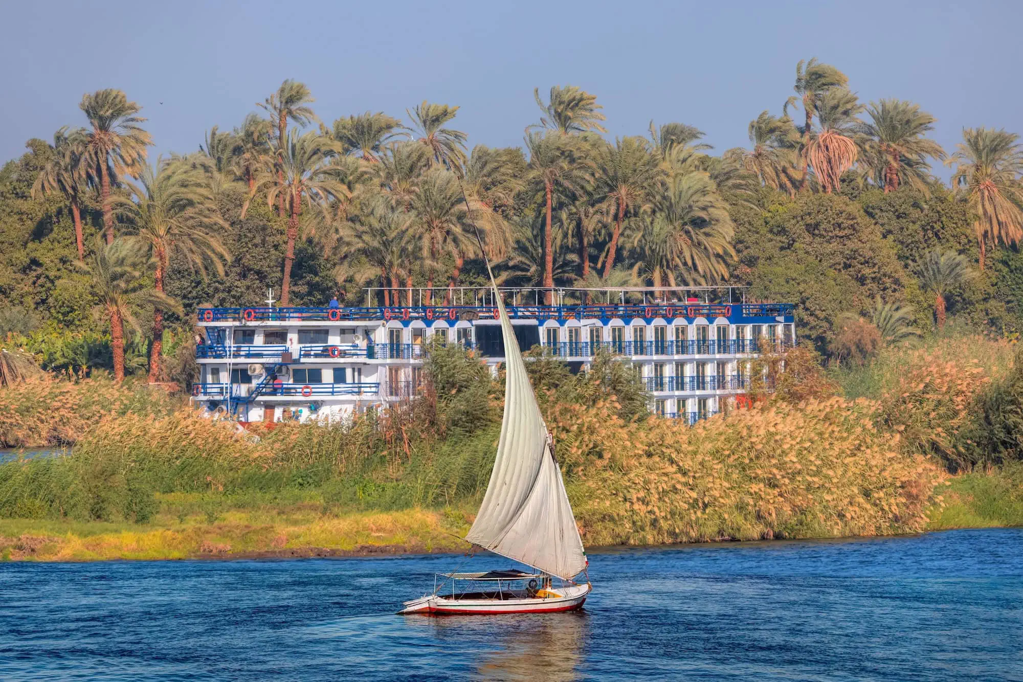 A traditional Egyptian felucca sailboat passing a multi-deck Luxury Nile Cruise vessel on the Nile River at Aswan, Egypt — the cruise is the standard Luxor–Aswan itinerary, the felucca is a half-day excursion in Aswan