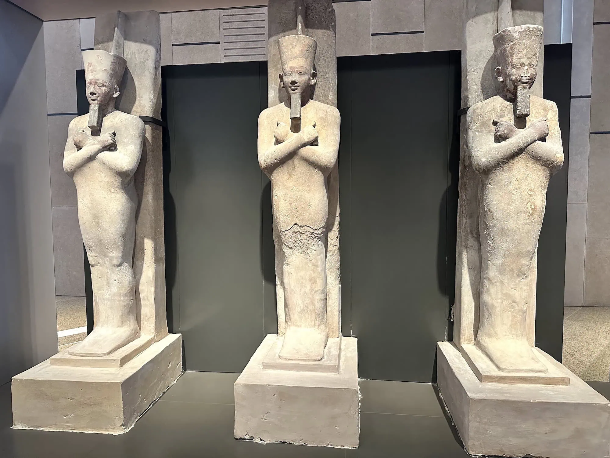 Three painted limestone Osiride statues of Pharaoh Senusret I (12th dynasty, c. 1971–1926 BC) on display at the Grand Egyptian Museum (GEM) in Giza, Egypt — depicting the king in mummiform pose with crook and flail, originally from his pyramid complex at El-Lisht