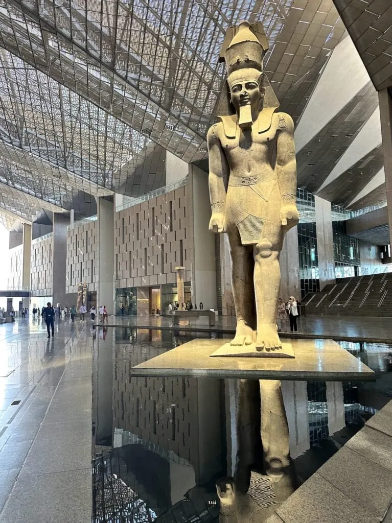 The Grand Hall atrium of the Grand Egyptian Museum, showing the towering 11-meter colossal statue of Ramses II as the central feature, with high ceilings and natural light from large windows.