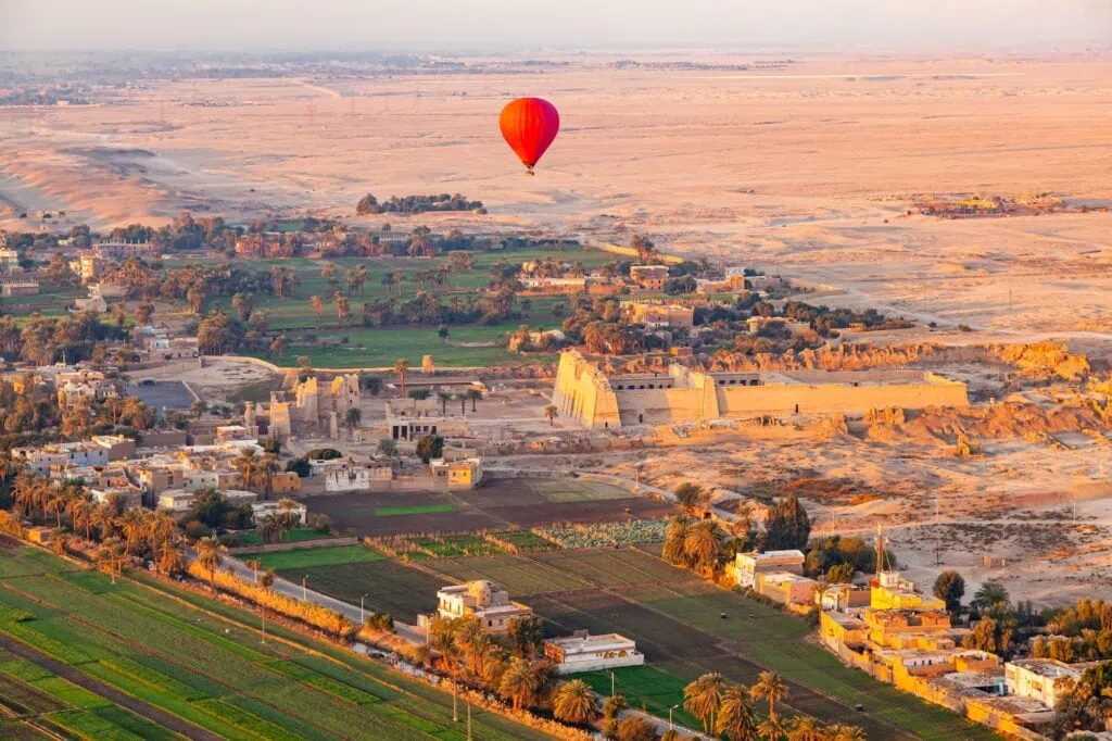 Hot-air balloons at sunrise over the Mortuary Temple of Ramses III at Medinet Habu on Luxor's West Bank, with the temple's intact pylons, courtyards, and surrounding mudbrick enclosure visible from the air.