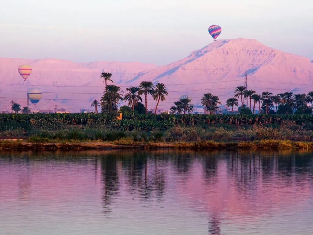 Hot air balloons rising at sunrise over Luxor's West Bank, with the Valley of the Kings, the Ramesseum, and the Colossi of Memnon in the desert landscape below, an optional pre-dawn add-on excursion on day two of the standard 4-day Luxor–Aswan Nile cruise itinerary.