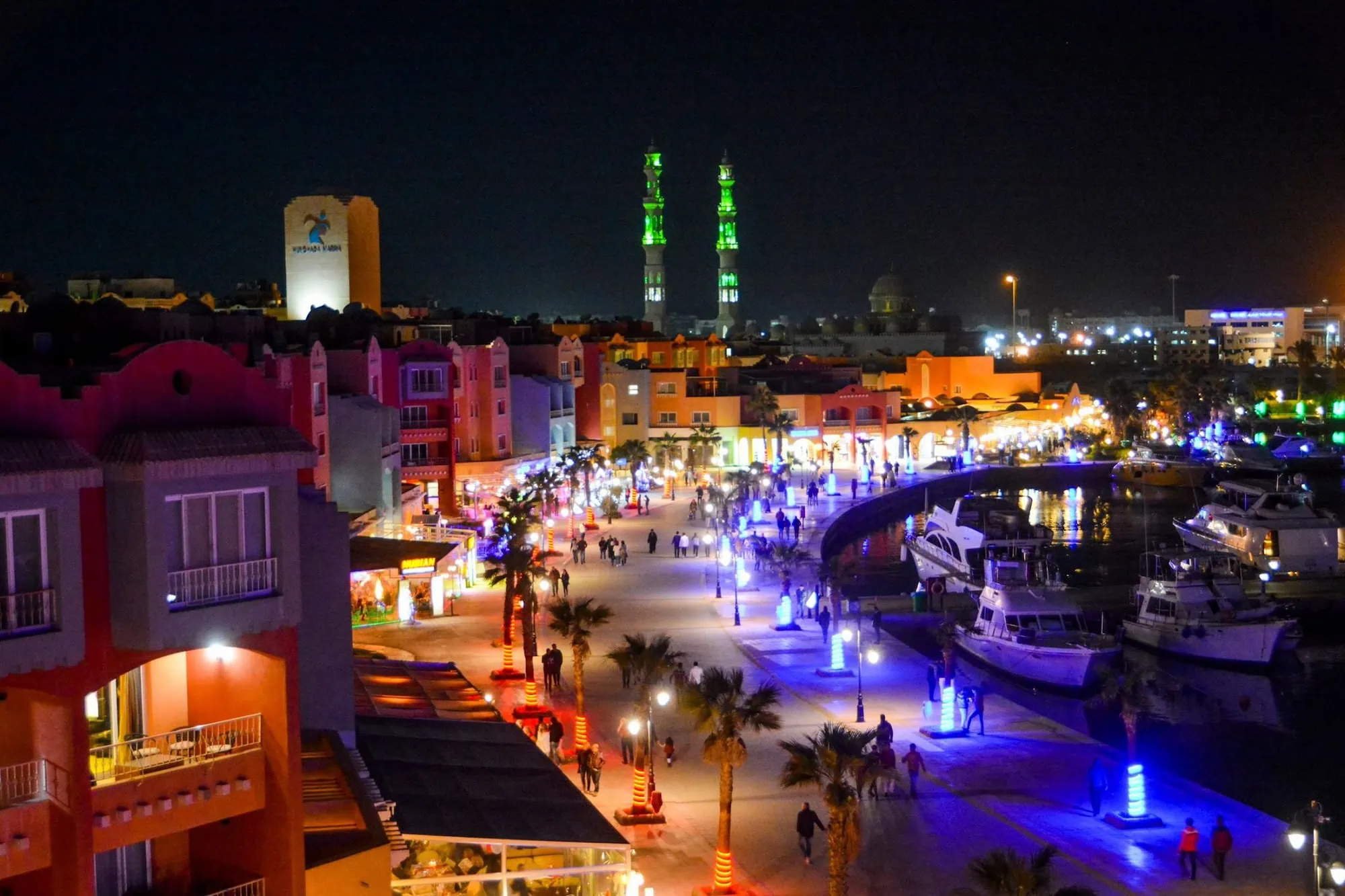 Hurghada New Marina by night with colorful waterfront buildings, moored yachts, and pedestrian promenade lit by evening lights