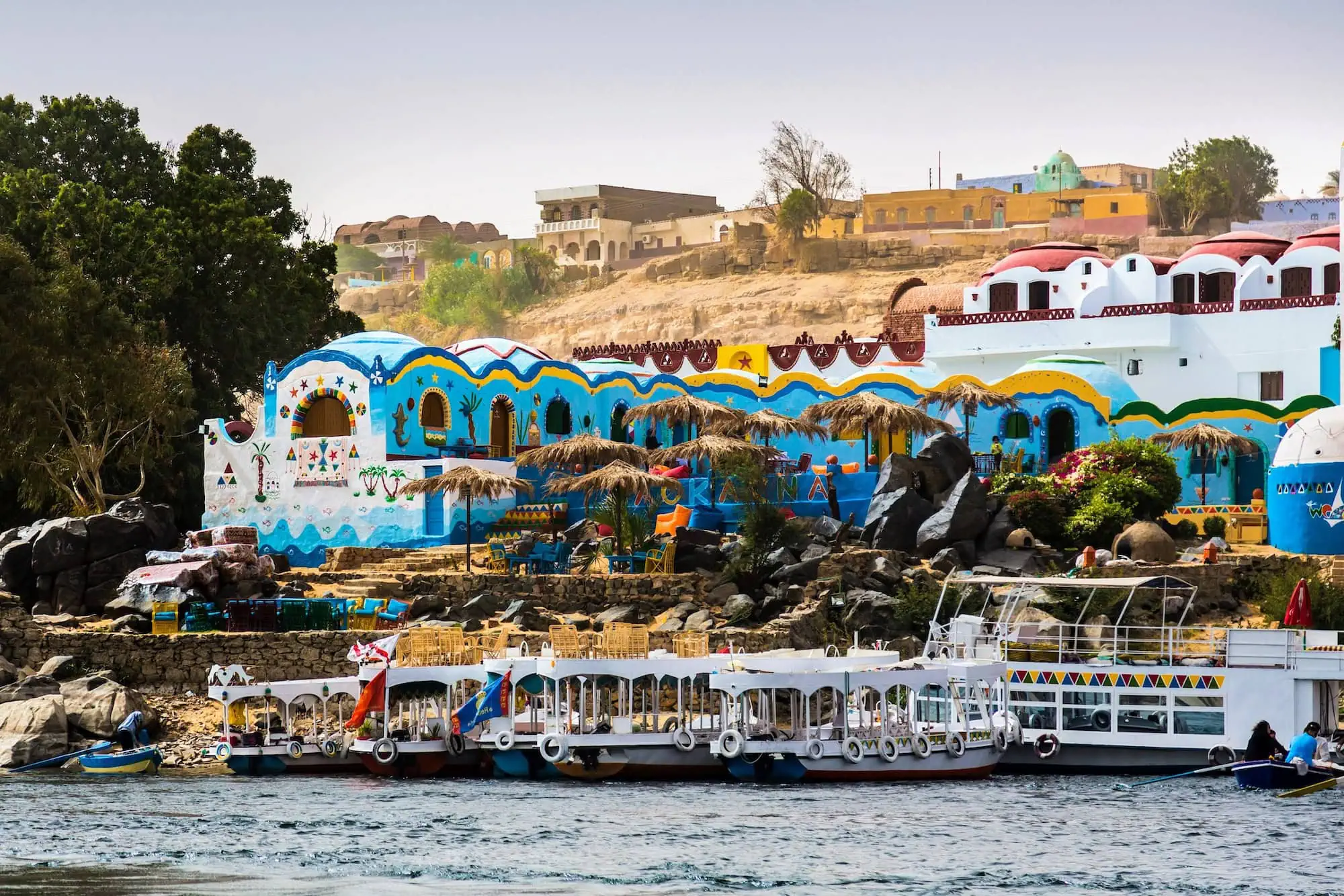 ourist boats on the Nile River in front of a Nubian village near Aswan, with colourful painted houses rising up the riverbank, Egypt.