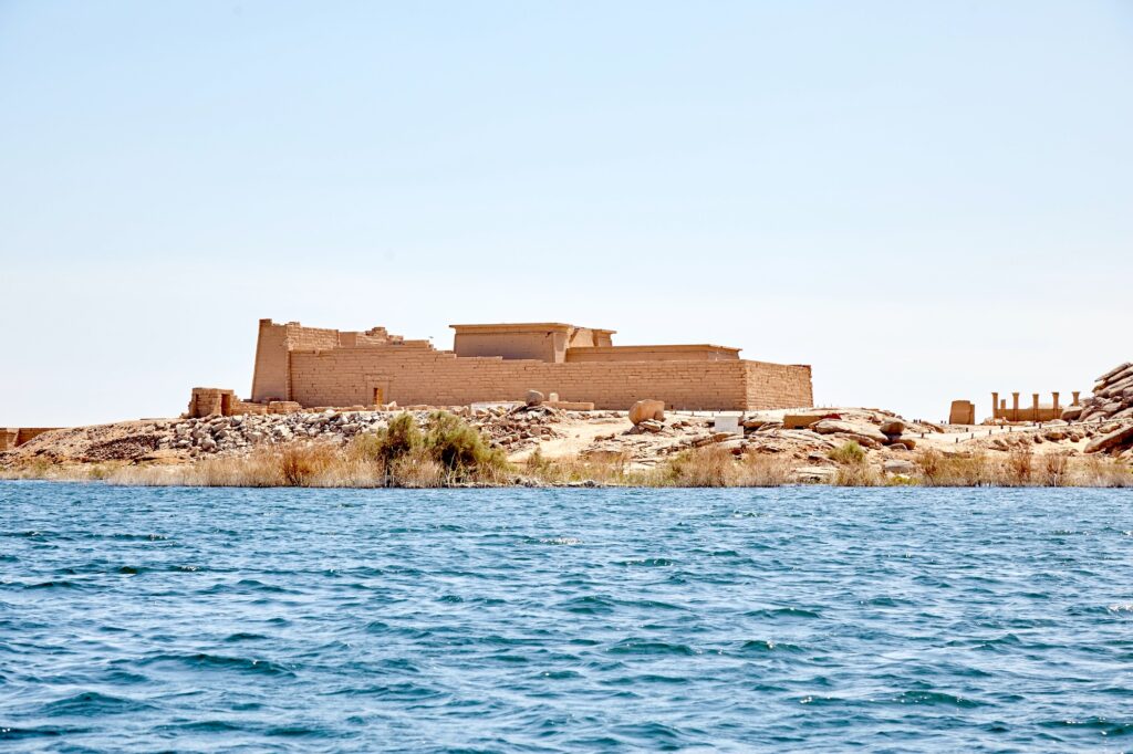 The sandstone walls and columns of the Temple of Kalabsha standing on the rocky shore of New Kalabsha, with the blue water of Lake Nasser visible beside the temple.