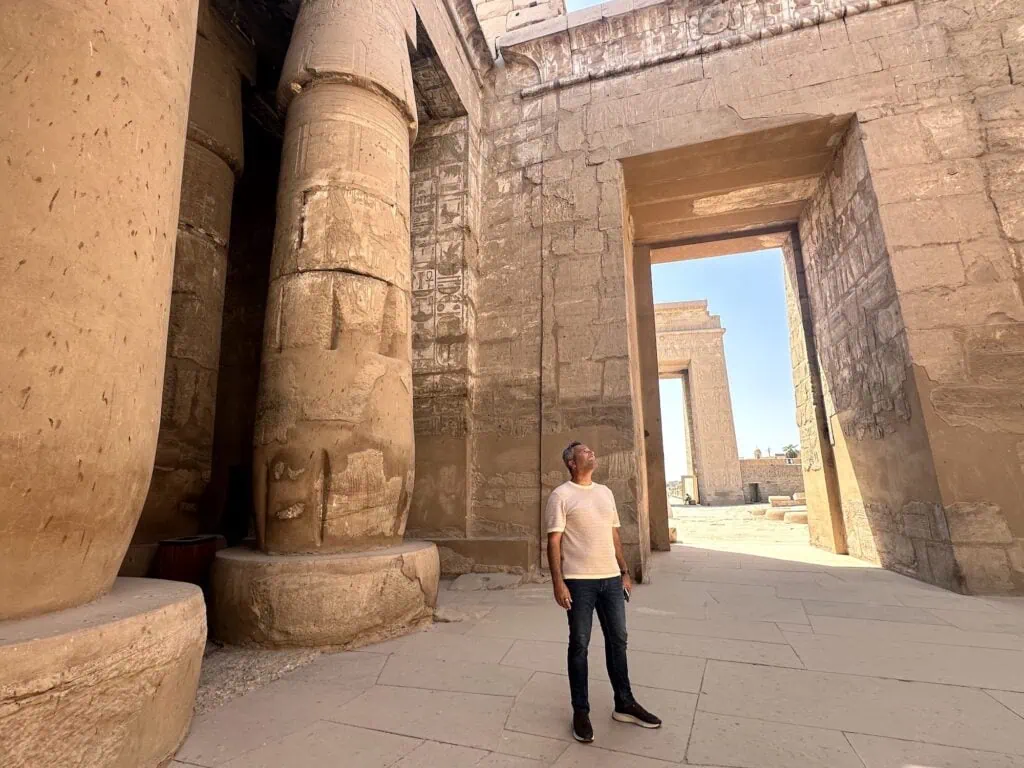 A visitor at Medinet Habu mortuary temple on the Luxor West Bank, Egypt — Ramses III's funerary temple from the 12th century BC, with massive sandstone columns and sunk-relief carvings depicting the pharaoh's battles against the Sea Peoples