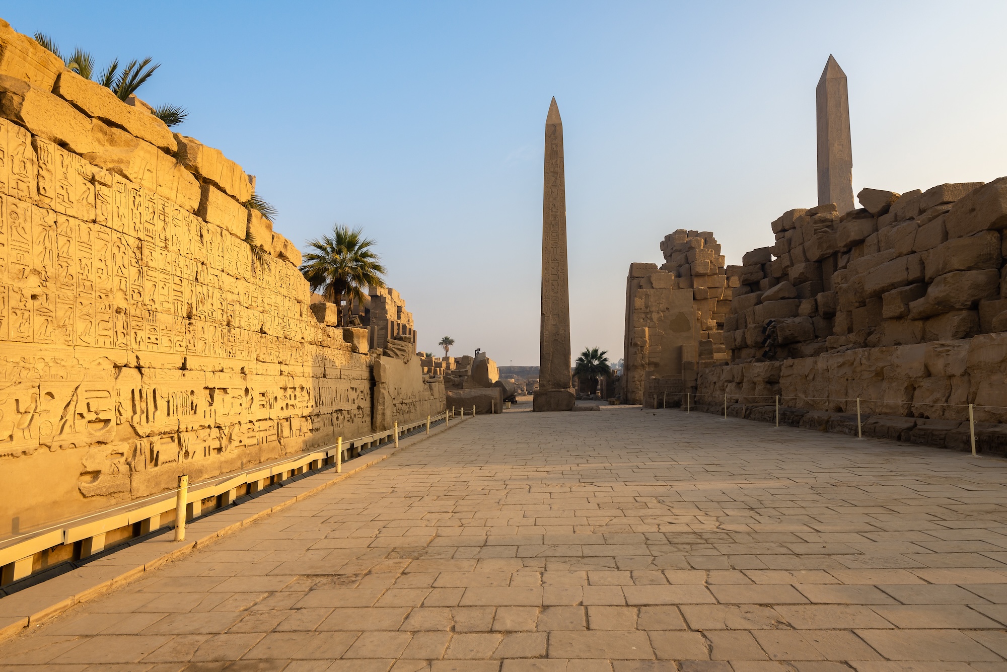 The Karnak Temple complex in Luxor, showing stone processional pathways, standing obelisks, massive pylons, and hieroglyph-covered walls under clear sky.
