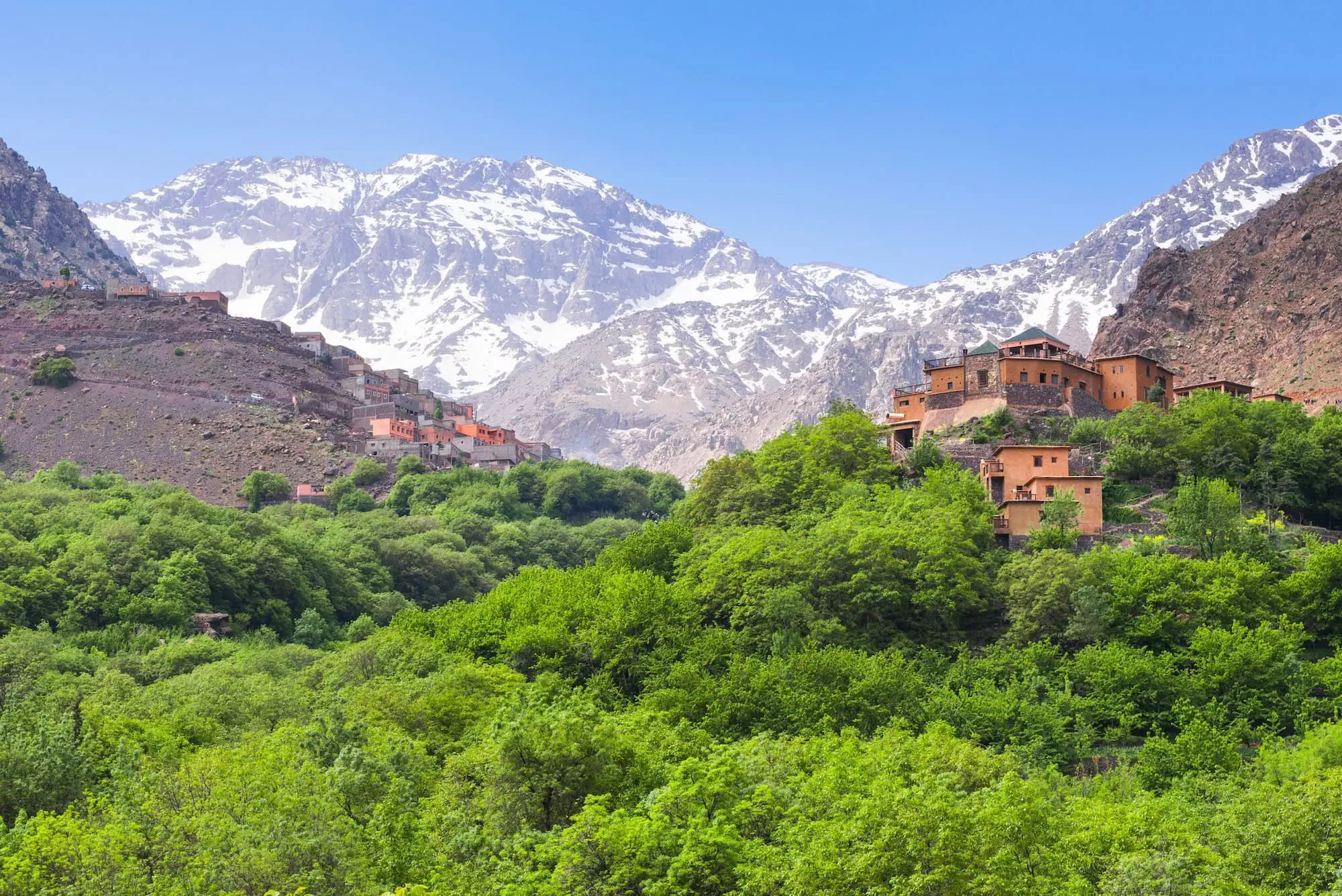 Traditional Berber village buildings in the Atlas Mountains with snow-capped peaks and green vegetation