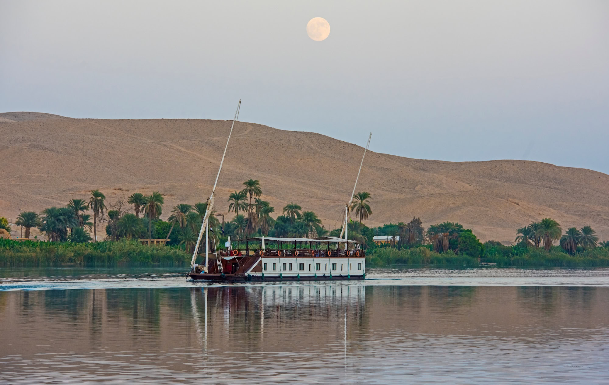 Traditional dahabiya boat sailing on the Nile River at sunset with desert cliffs and palm trees