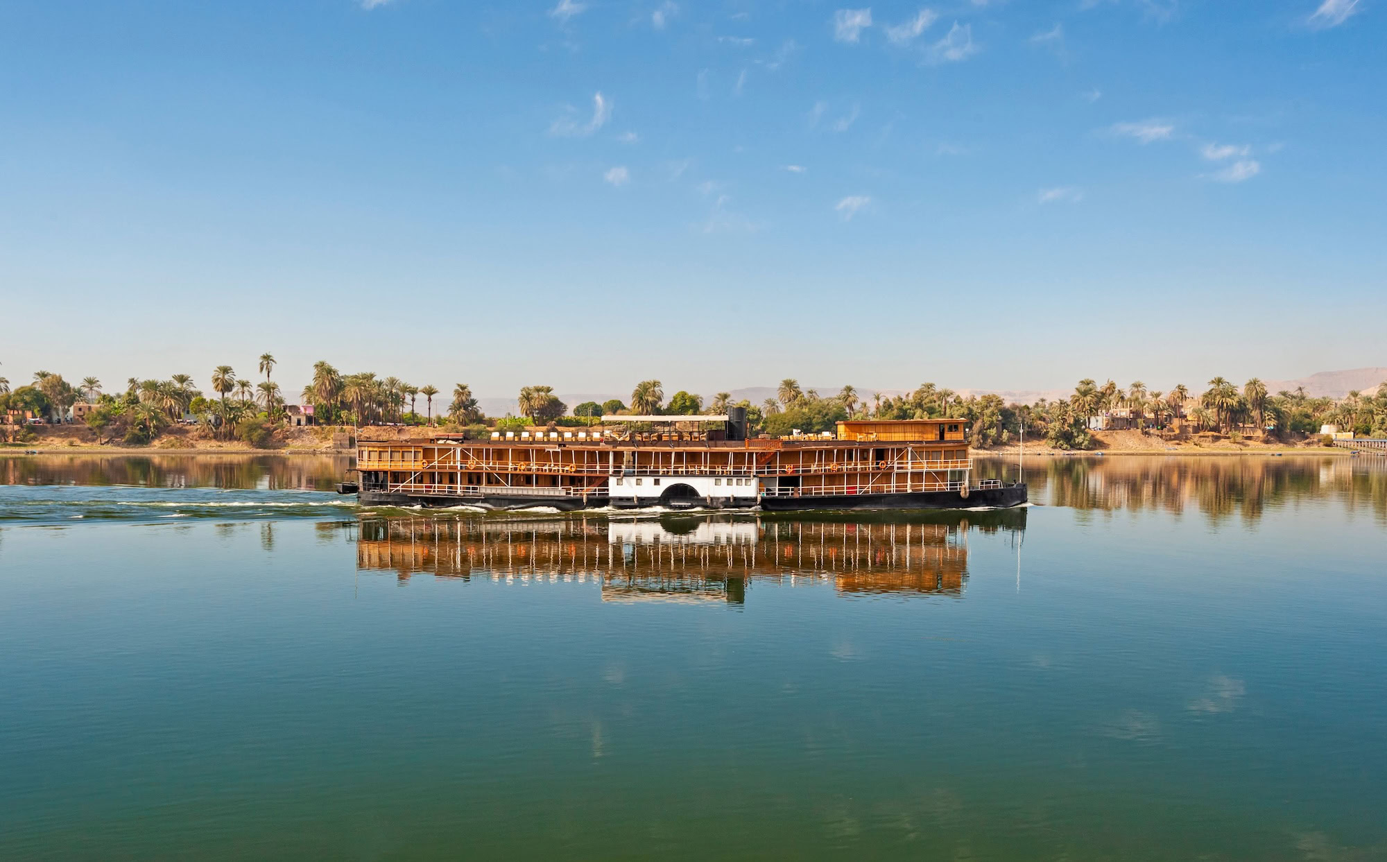 Nile River cruise boat sailing on calm waters with palm trees along the shoreline