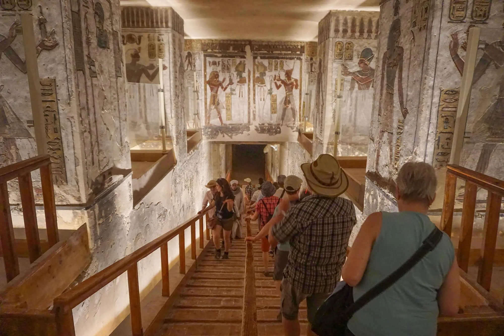 Travelers descending wooden access stairs into the entrance corridor of an ancient Egyptian tomb in the Valley of the Kings, with hieroglyphic murals visible on the walls