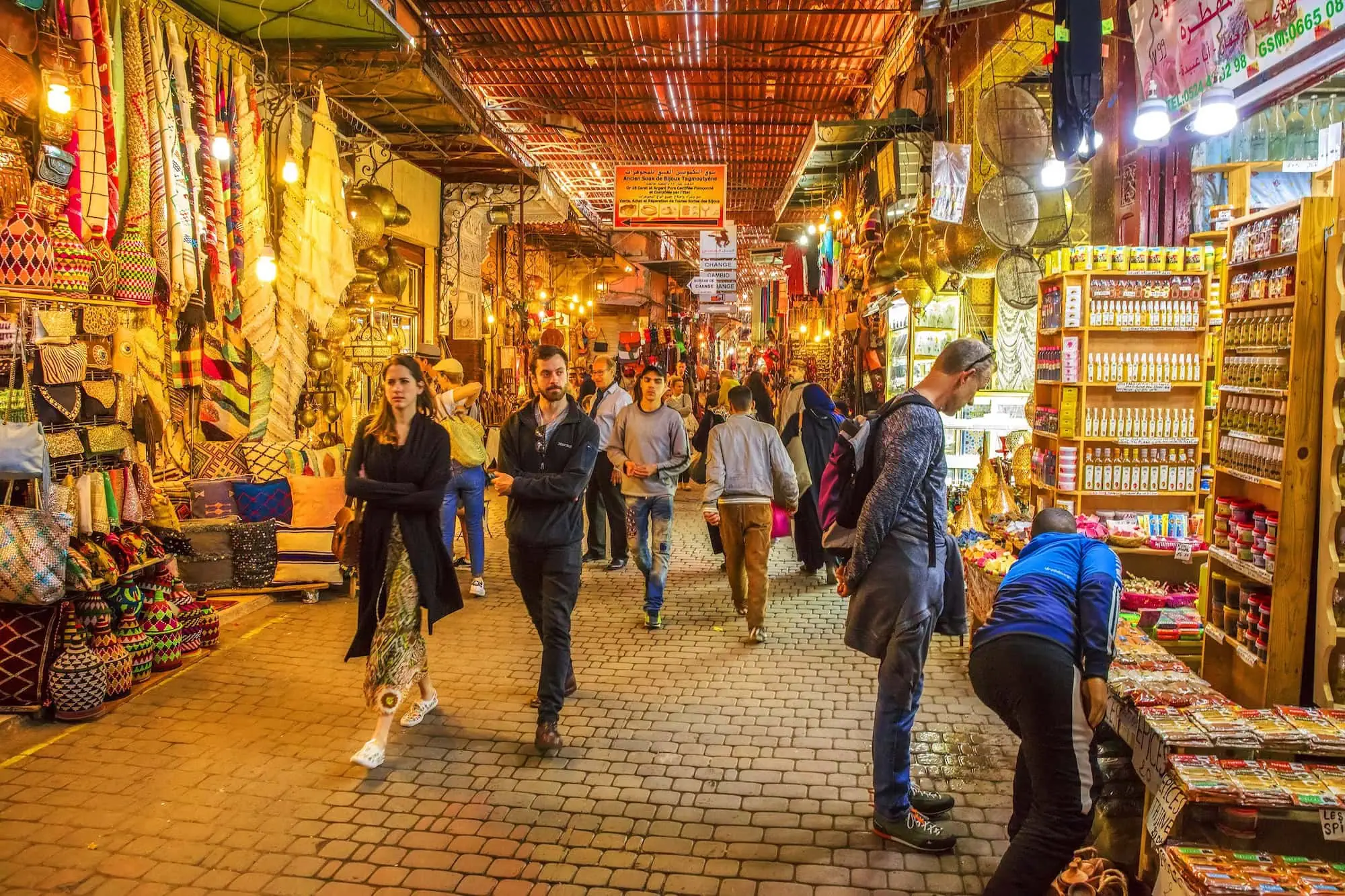 Bustling traditional Marrakech souk with tourists and locals browsing colorful textiles and goods in covered walkways