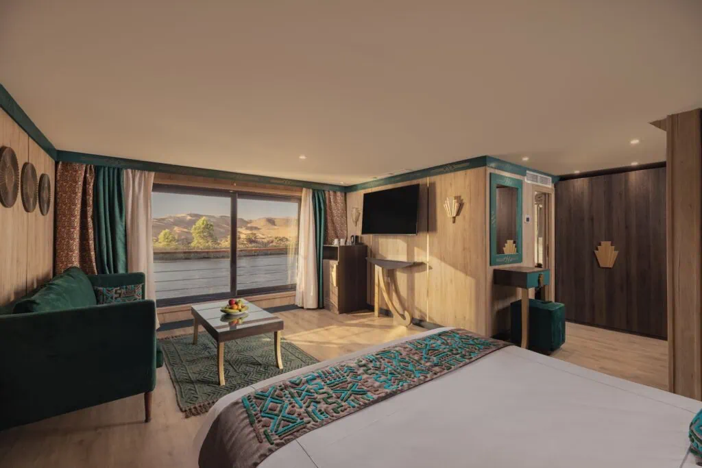 The interior of a luxury suite cabin aboard a 5-star Nile cruise vessel, with a bed, a separate seating area, and large panoramic windows looking onto the Nile.