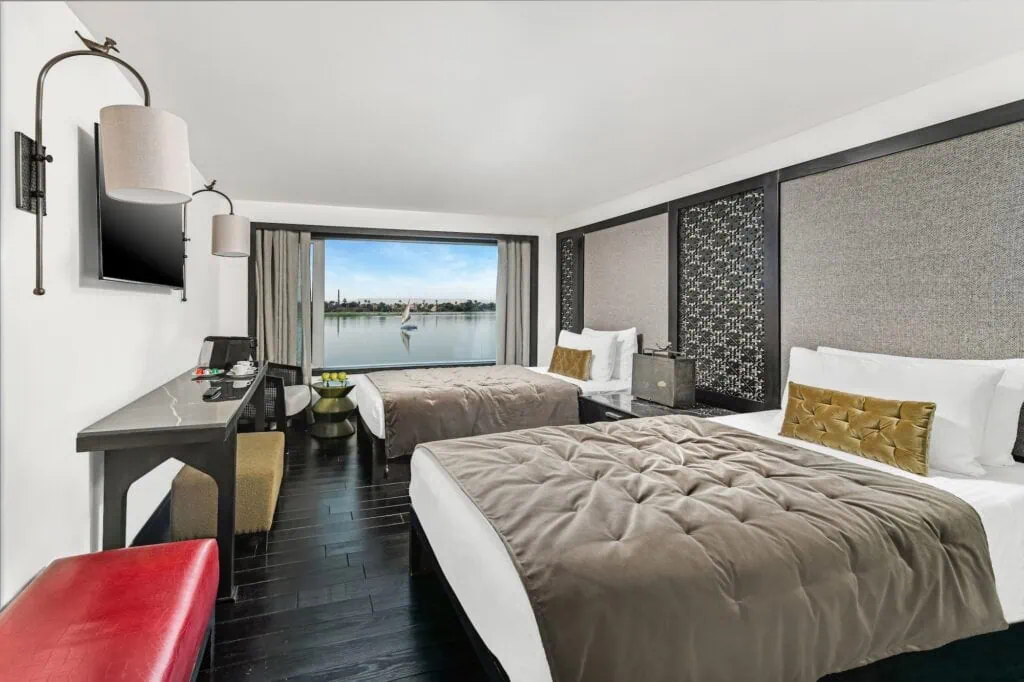 The interior of a cabin aboard the Mövenpick MS Darakum, with a bed, panoramic window, and a wide view of the Nile River from inside the cabin.