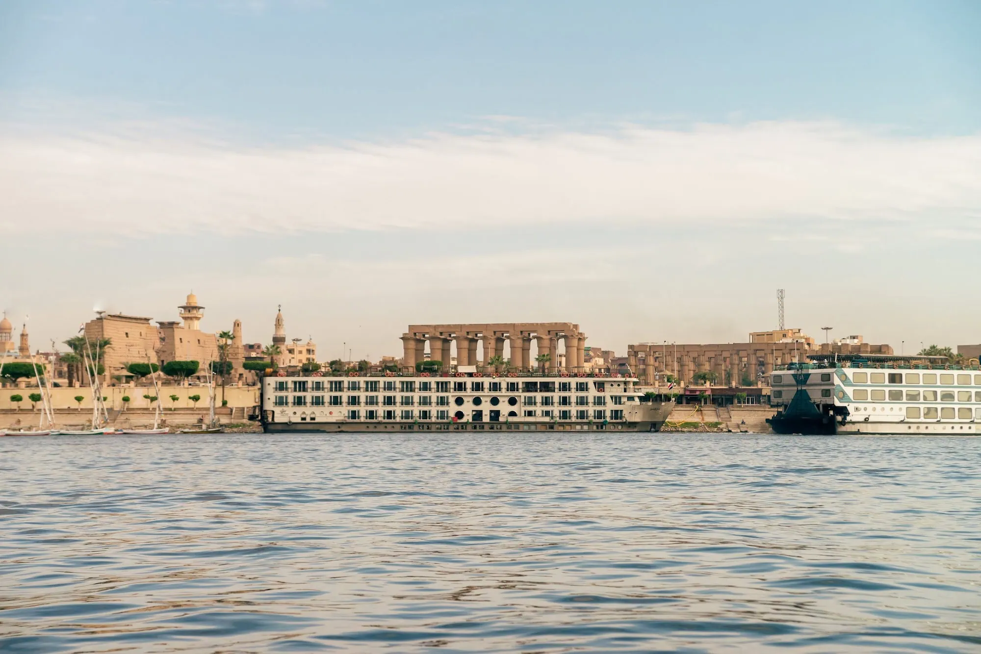A row of Luxury Nile Cruise vessels moored along the east bank of the Nile in Luxor, with city buildings rising behind