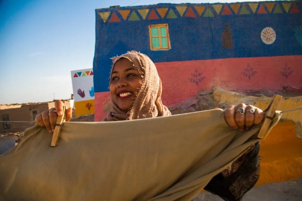 A young Nubian woman in patterned dress hanging laundry between brightly painted village houses