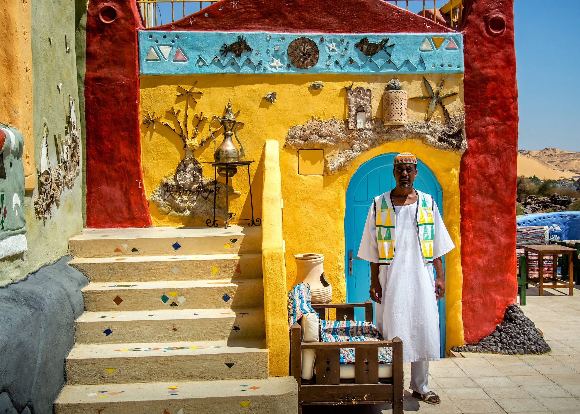 Man in traditional Nubian dress sitting by colorful house with pottery and crafts