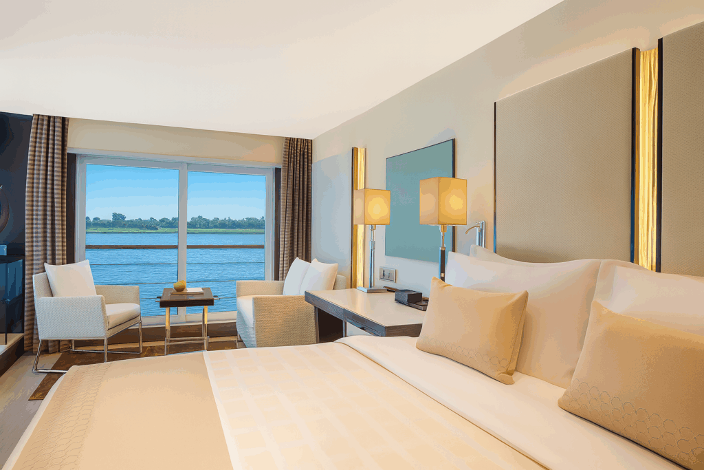 The interior of a cabin aboard a 5-star Nile cruise vessel, with a made bed, wooden detailing, and a large panoramic window looking out onto the Nile.