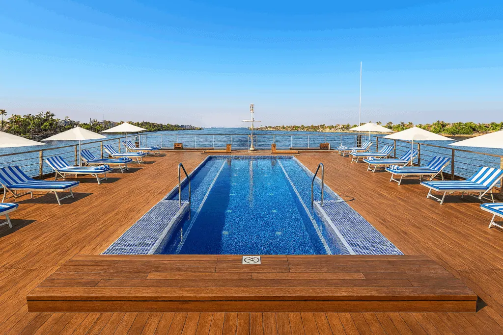 The pool deck of a 5-star Nile cruise vessel with lounge chairs arranged around the pool, overlooking the Nile River and its banks under clear sky.