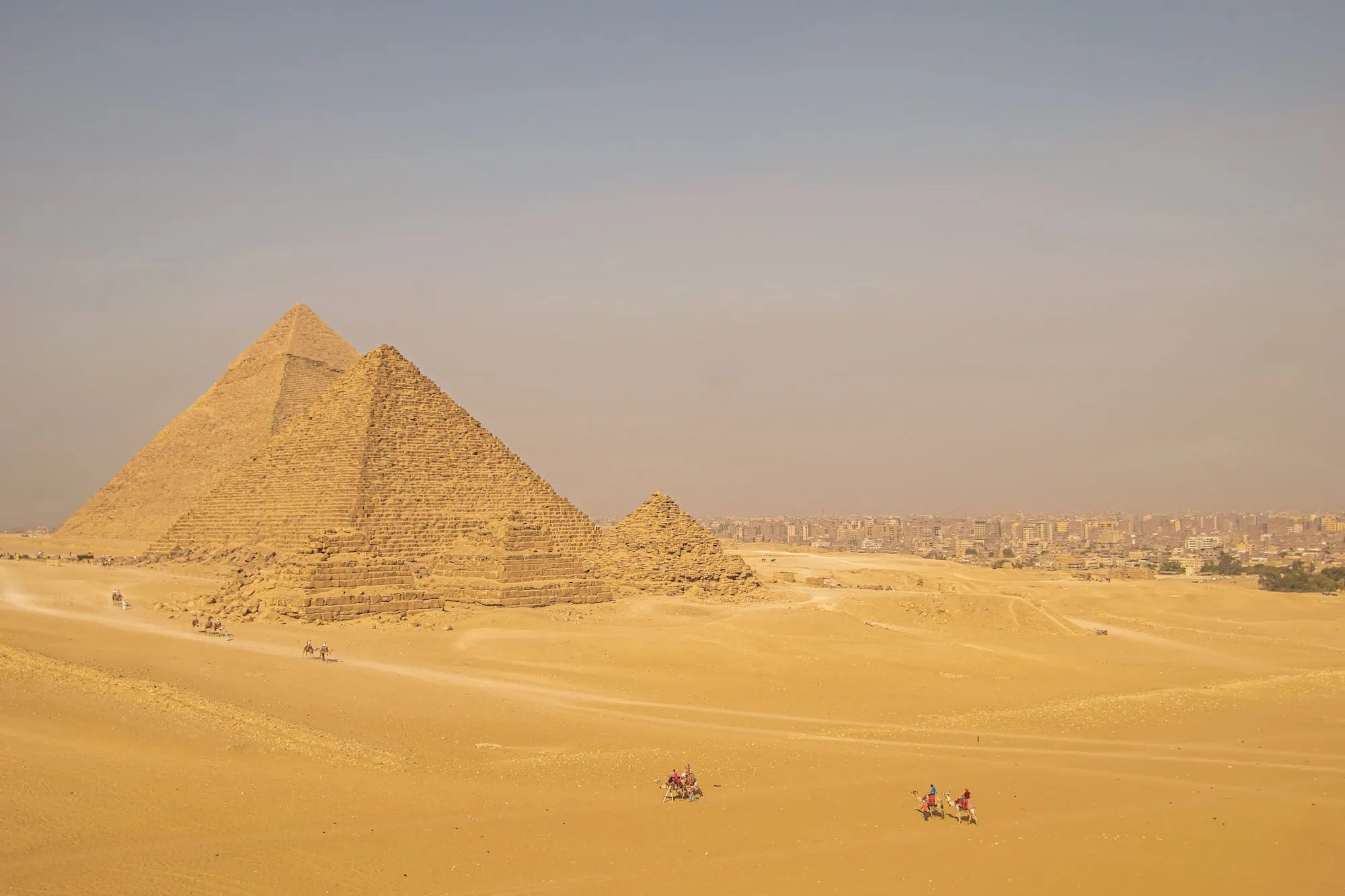Panoramic view of the Giza pyramid plateau outside Cairo, with small groups of tourists and camels in the foreground and the pyramids of Khufu, Khafre, and Menkaure behind