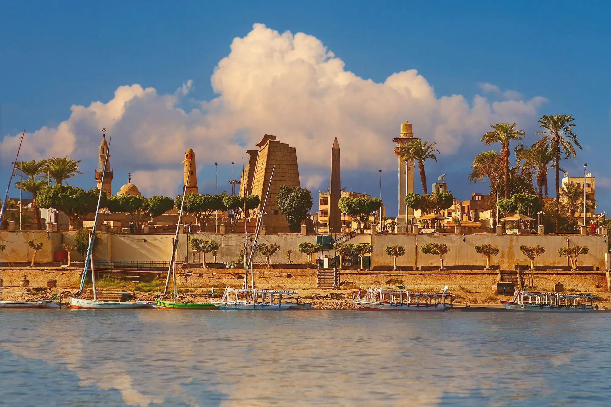 Traditional Egyptian felucca sailboats on the Nile River at sunset with Luxor temples and palm trees in background