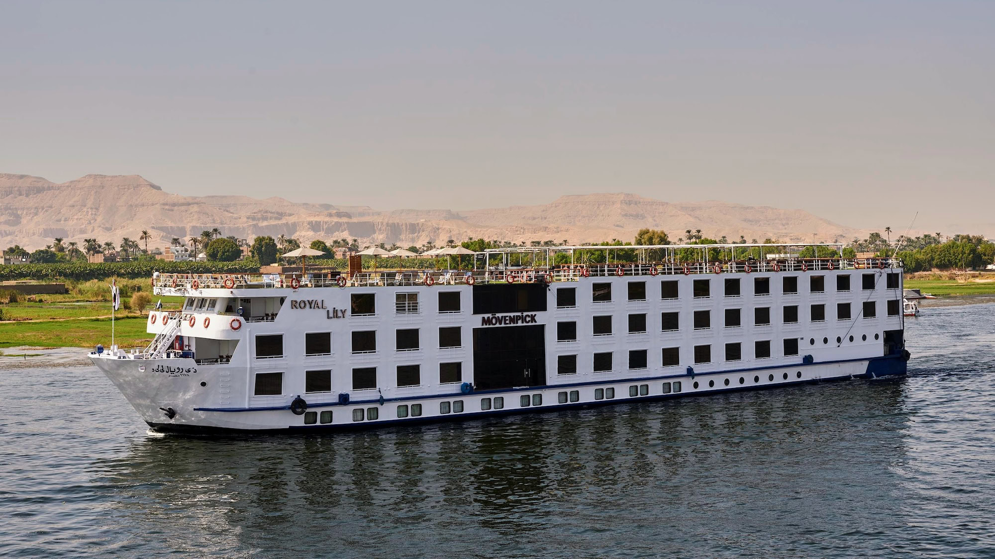 Luxury cruise ship sailing on the Nile River past desert mountains and palm trees