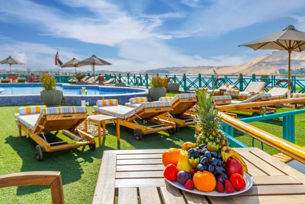 A plate of sliced fresh fruit beside sun loungers and a parasol on the upper deck of a Luxury Nile Cruise vessel, with the Nile and palm-lined banks visible behind.