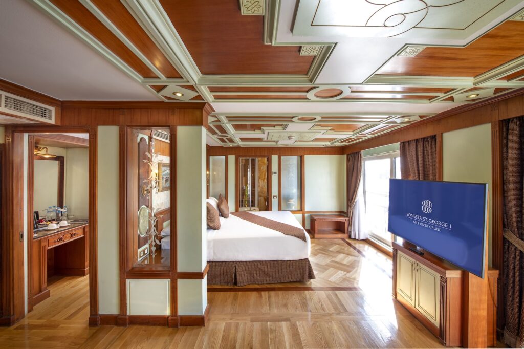 Royal Suite bedroom aboard the Sonesta St. George Luxury Nile Cruise vessel, with floor-to-ceiling river-facing windows, dark-wood classic furnishings, and a private sitting area