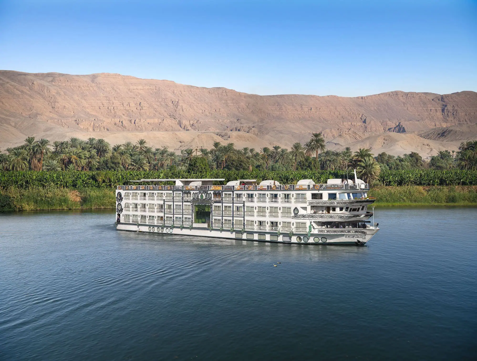 A white multi-deck 5-star Nile cruise vessel sailing on the blue water of the Nile, with palm trees along the bank and desert mountains rising behind.