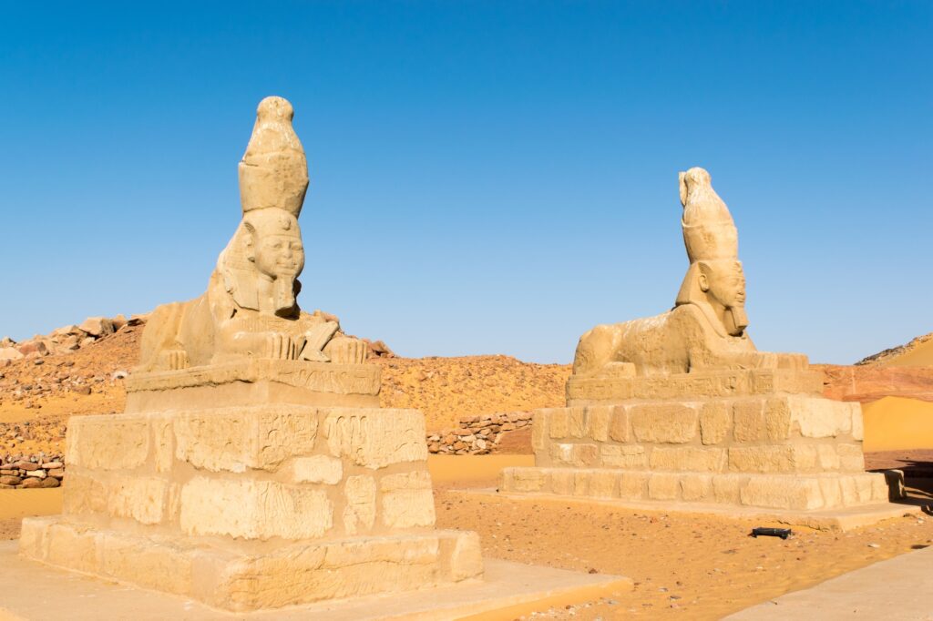 A weathered stone sphinx standing on the entrance avenue of Wadi el-Seboua temple in Nubia, with the desert landscape of the Lake Nasser region behind it.