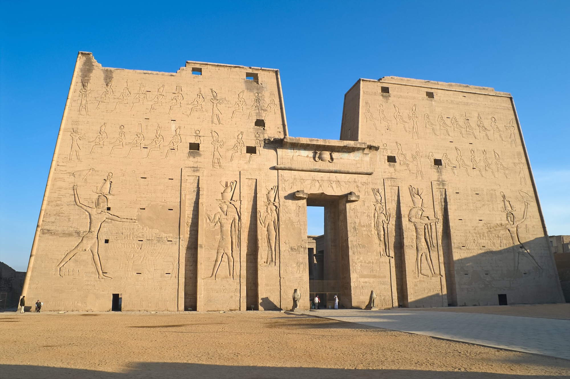 The massive entrance pylon and sandstone facade of the Ptolemaic Temple of Horus at Edfu, photographed from the temple forecourt, showing the deep-relief carvings of pharaoh and Horus the falcon god.