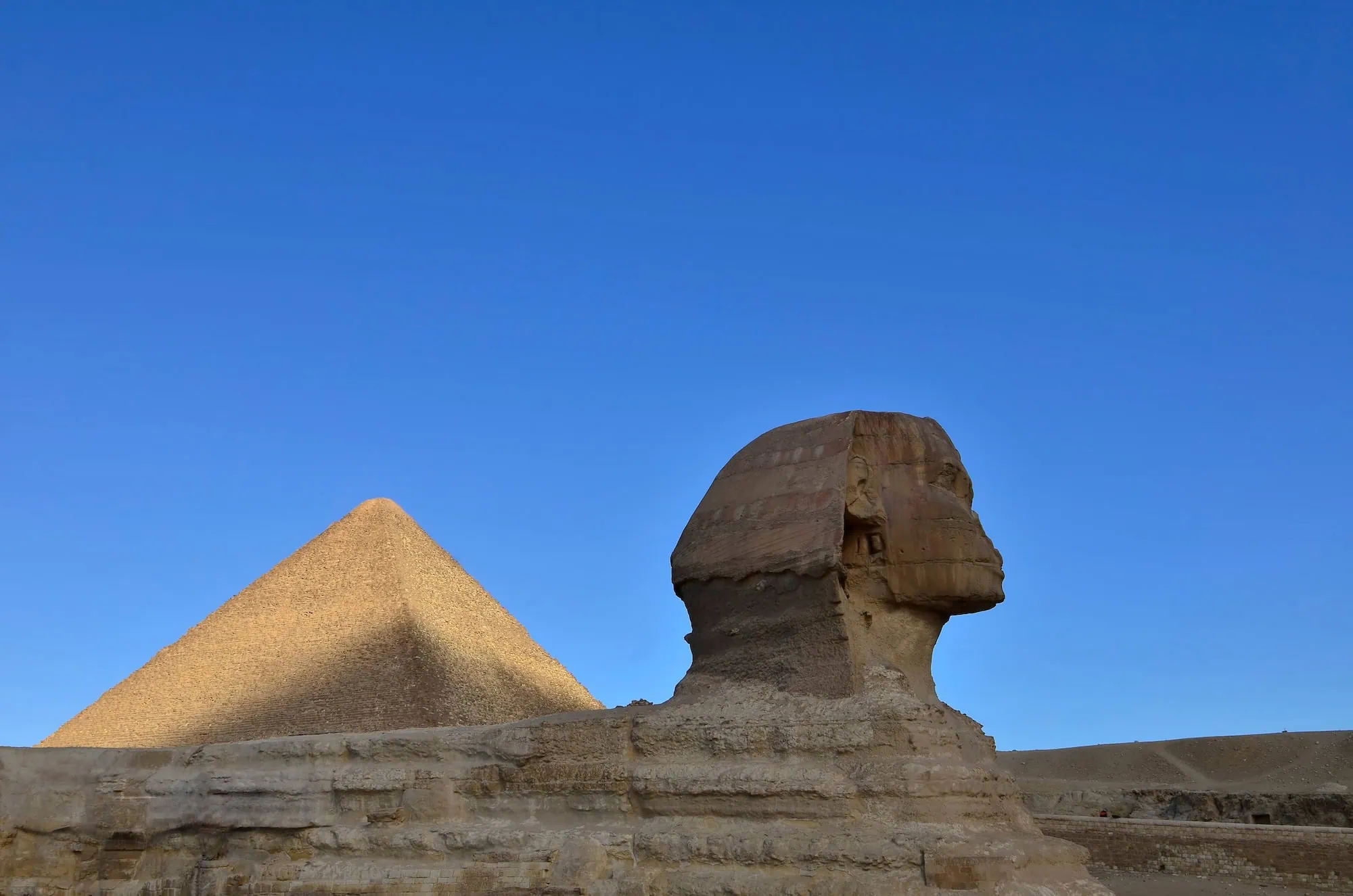 The Great Sphinx of Giza in the foreground with the Great Pyramid of Khufu behind it under a bright blue sky on the Giza plateau, Egypt.