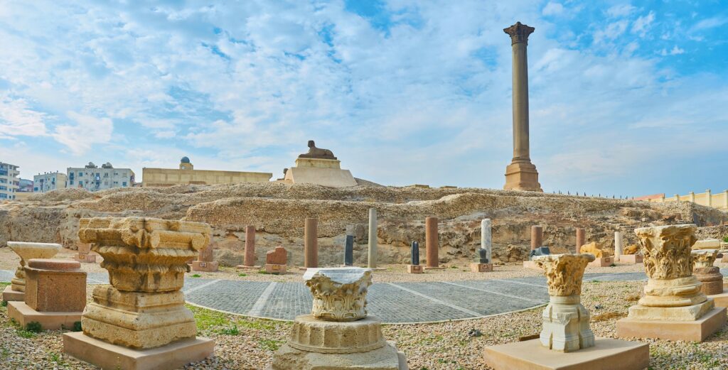 Pompey's Pillar rising behind a sphinx statue with surrounding ruins of the Serapeum Temple at Amoud Al Sawari archaeological site, Alexandria