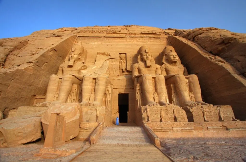 The facade of the Great Temple of Ramses II at Abu Simbel, showing four colossal seated statues of the pharaoh carved into the sandstone cliff under a clear blue sky. Lake Nasser Cruises