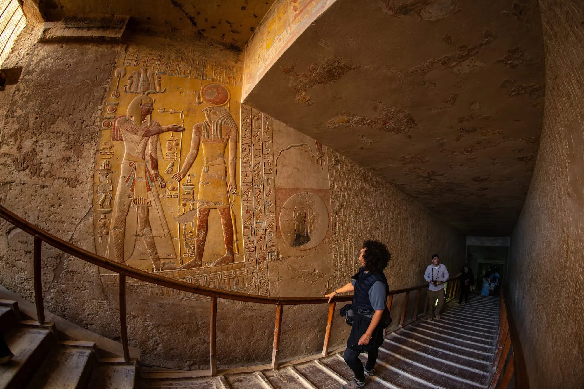 Visitors descending a long sloping staircase inside a royal tomb in the Valley of the Kings, past a vivid painted relief of a pharaoh and a sun god, Luxor, Egypt.